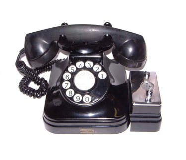 Automatic central battery operated telephone set. It has a token box to convert it into a public telephone. Model made of black bakelite with quadrangular base and truncated pyramid-shaped top. The fork has a very open U-shape, with a central battery....